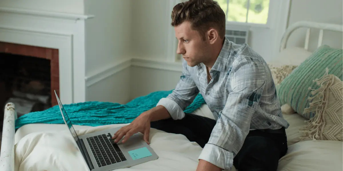 Man researching mattresses on his Mac, sitting on a full bed with white blankets and green pillows.